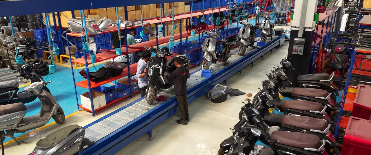 Mercury electric scooters lined up along the final assembly line, technicians fitting bodywork at the bench stations beside them.