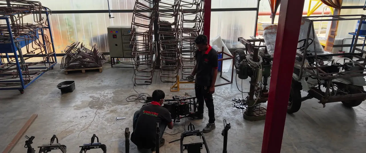 Two Mercury technicians welding an electric three-wheeler chassis frame on a jig, finished frames stacked behind them.